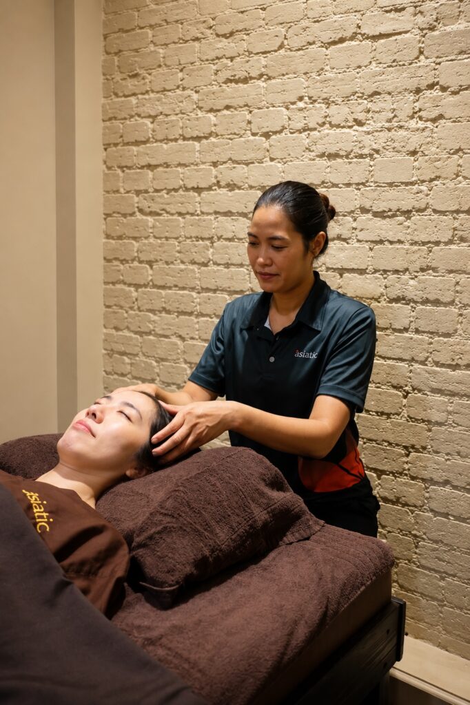 Asiatic massage therapist performing gentle head massage in a calm brick-walled treatment room in London.