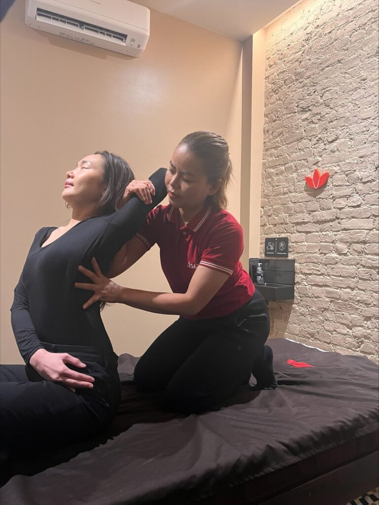 Asiatic massage therapist gently stretching the back of a client. The therapist is using their hands to apply pressure and gently pull the client's body into a stretch. 